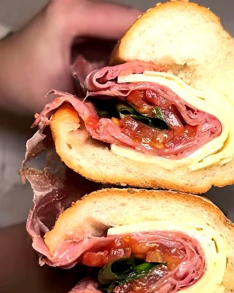 An Italian-style hoagie on a seeded roll, cut in half: sliced meats, cheese, tomato and greens