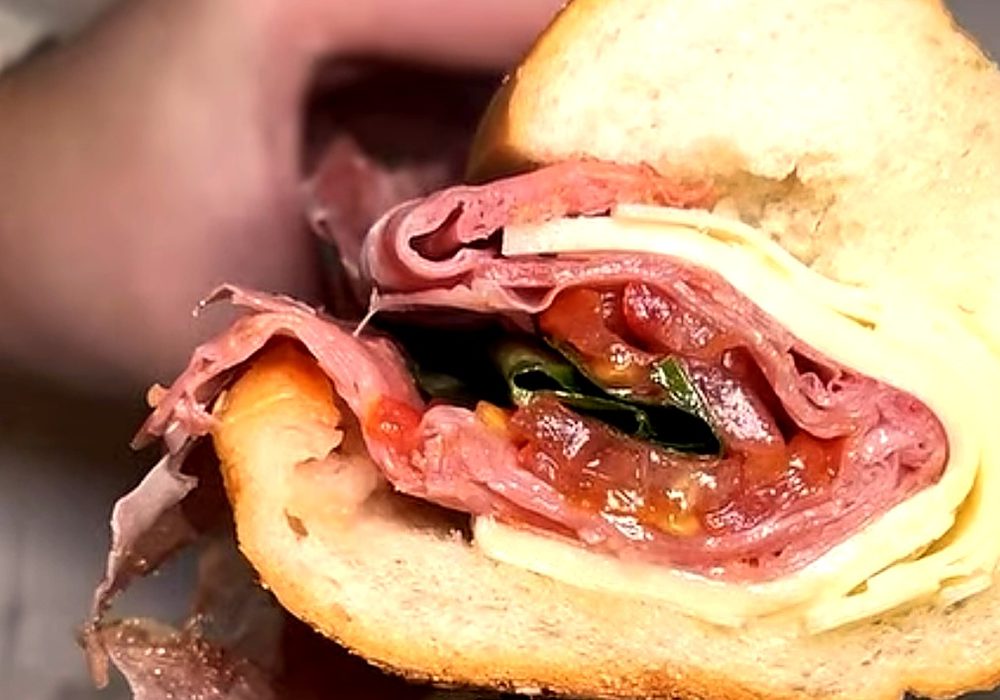 An Italian-style hoagie on a seeded roll, cut in half: sliced meats, cheese, tomato and greens